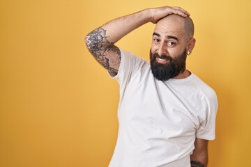 Young hispanic man with beard and tattoos standing over yellow background smiling confident touching hair with hand up gesture, posing attractive and fashionable