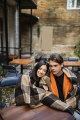 Cheerful asian woman in blanket hugging boyfriend on terrace of cafe.