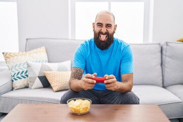 Young hispanic man with beard and tattoos playing video game sitting on the sofa sticking tongue out happy with funny expression.