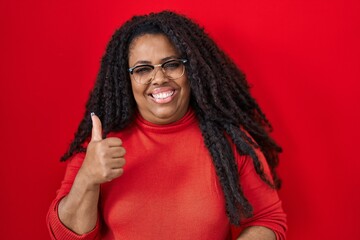 Plus size hispanic woman standing over red background doing happy thumbs up gesture with hand. approving expression looking at the camera showing success.