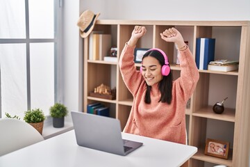 Young hispanic woman listening to music and dancing sitting on table at home