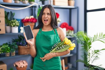 Brunette woman working at florist shop holding smartphone angry and mad screaming frustrated and...