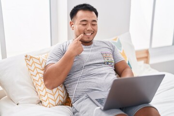 Chinese young man using laptop sitting on the bed smiling happy pointing with hand and finger