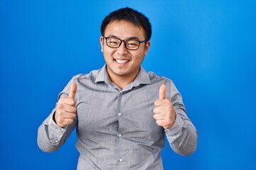 Young chinese man standing over blue background success sign doing positive gesture with hand, thumbs up smiling and happy. cheerful expression and winner gesture.