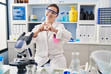 Hispanic girl with down syndrome working at scientist laboratory smiling in love doing heart symbol shape with hands. romantic concept.