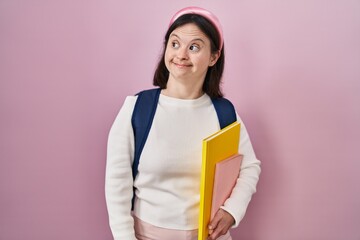 Woman with down syndrome wearing student backpack and holding books smiling looking to the side and staring away thinking.