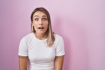 Blonde caucasian woman standing over pink background afraid and shocked with surprise and amazed expression, fear and excited face.