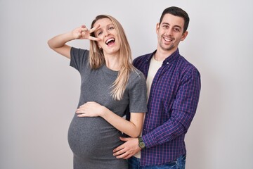 Young couple expecting a baby standing over white background doing peace symbol with fingers over face, smiling cheerful showing victory