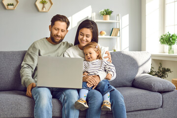 Mom, dad and their little daughter are relaxing at home watching different videos on laptop. Cheerful young Caucasian woman man and two-year-old girl sitting together on sofa in living room.