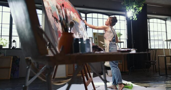 A young caucasian woman paints a picture with a wide flat brush in a large fashion workshop. Woman artist paints an abstract picture. Contemporary female artist creating abstract contemporary art