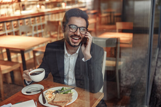 Smiling indian businessman is talking phone with client during lunch time in cozy cafe