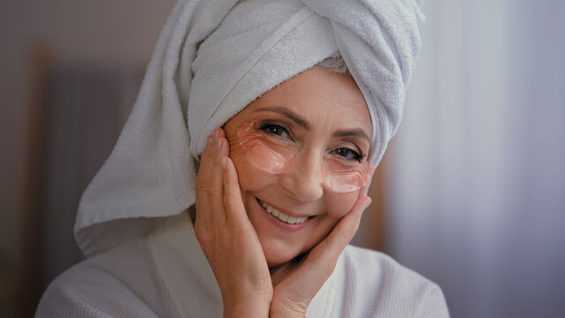 Positive portrait happy senior lady with anti wrinkle skin hydrogel collagen patches under eyes smiling to camera old 60s Caucasian woman with towel on head enjoy morning beauty rituals with skincare 