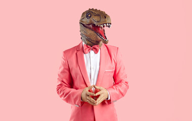 Portrait of a funny bizarre man wearing a goofy rubber dinosaur mask and a trendy pink suit with a...
