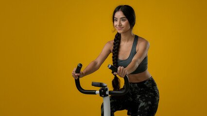 Ethnic Indian sexy strong slim slender woman cycling in yellow studio background cardio workout warming with sport stationary bicycle bike equipment biking female cyclist lady sports enjoy exercise