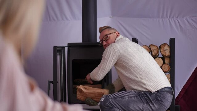 Confident man putting firewood closing furnace talking to blurred woman smiling. Portrait of Caucasian husband chatting with wife resting in eco hotel room. Lifestyle and leisure