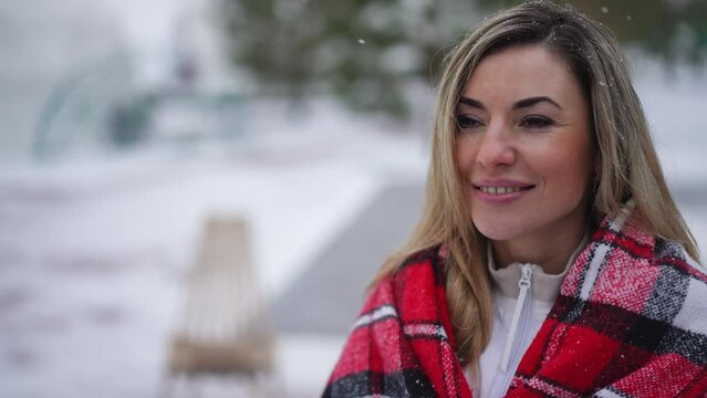 Live camera zoom in to smiling confident woman wrapping in blanket standing on winter day outdoors with white snow falling. Portrait of happy relaxed Caucasian beautiful lady enjoying vacations
