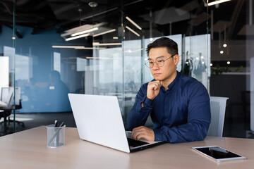 Serious thinking asian businessman working inside modern office, mature man in shirt and glasses...