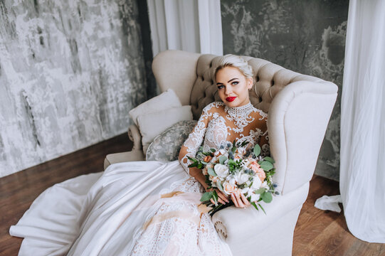 A beautiful blonde bride in a long lace dress sits on a sofa in the studio, indoors with a bouquet of flowers in her hands on a gray background. Wedding photography, portrait.