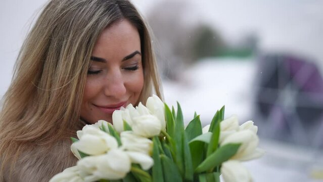 Close-up portrait of smiling satisfied Caucasian woman posing with bouquet of white tulips on Valentine's Day outdoors. Happy beautiful lady looking at camera standing with flowers as snow falling