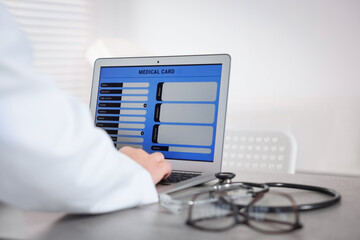 Doctor using laptop to fill out patient's medical card at table in clinic, closeup