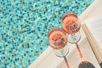 Keuken foto achterwand Tasty glasses of rose wine and open book on swimming pool edge, space for text © New Africa