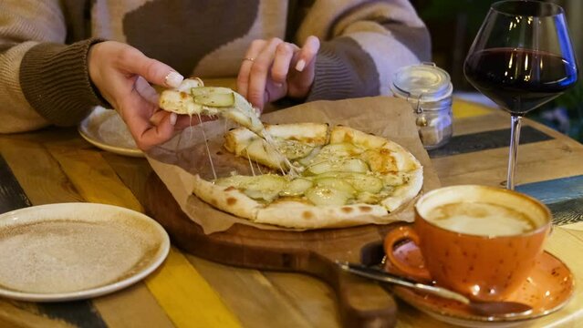 A woman's hand takes a piece of hot pizza with pear and cheese, sitting in a cafe in the evening.