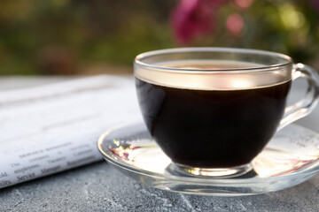 Glass cup with coffee and newspaper on grey table outdoors, closeup. Morning ritual