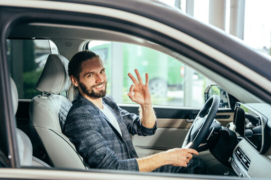 Happy male owner of new car auto gesturing sign ok on camera. Handsome man sitting inside modern car and feeling happiness from expensive purchase.