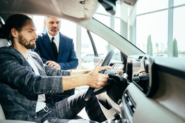 Caucasian male client customer man sitting at the wheel of new car doing test-drive before buying auto while male shop assistant helping him choose.