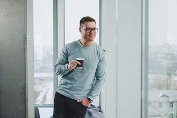 Smiling man in casual clothes standing near window with coffee in modern work area with big window during daytime.