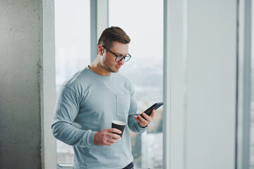 Smiling man in casual clothes standing near window with coffee and looking at smartphone in modern workspace with big window during daytime