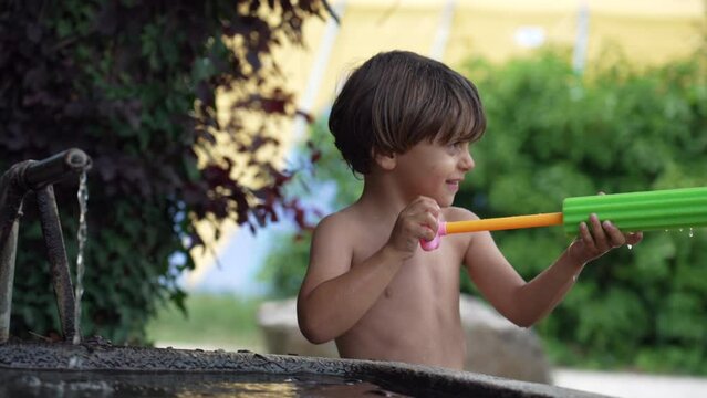 One happy child spraying water with foam toy blaster at outdoor backyard in summer day. Kid playing with water shirtless. Happiness concept