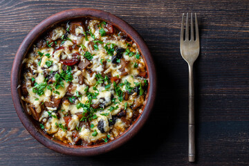 Stewed potato, carrot, onion, tomato, prunes and cheese in a clay bowl on wooden background, closeup, top view