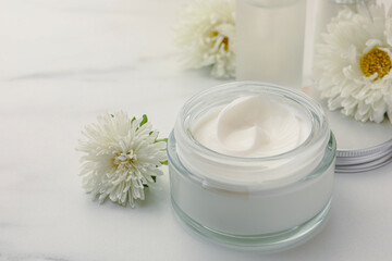 Fototapeta na wymiar Glass jar of face cream and flowers on white marble table. Space for text