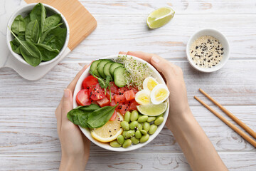Woman holding delicious poke bowl with quail eggs, fish and edamame beans at white wooden table, top view