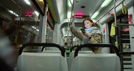 Young beautiful happy woman taking ride in bus standing and holding handrail. Attractive caucasian adult girl wearing coat and scarf looking at smartphone smiling riding in empty public transport.