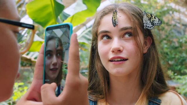 Friends, butterfly and taking pictures at zoo with phone for social media, happy memory or internet post. Mobile, tech and girl taking photos of insect on face of teenager while learning about bugs.