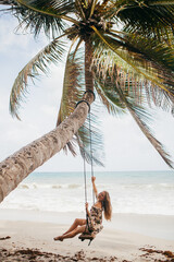 The girl on the beach rides on a swing.  Tropics, enjoying nature. Swing tied to a palm tree by the ocean.