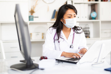 Experienced female physician in protective mask filling up medical forms on laptop while sitting at table in office