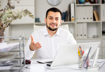 Portrait of positive man office worker sitting at table and making hand gesture. Businessman gesturing, looking at camera.