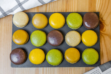 Green, orange, yellow and brown macarons on a slate on a wooden table with decorative cloth and newspaper
