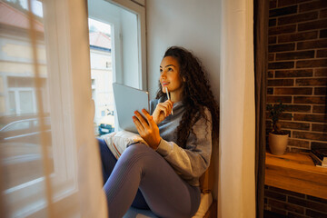 Young student sitting in a bay window, thinking about project ideas and making plans on her tablet