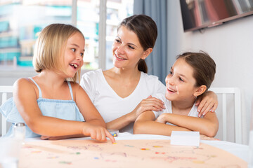 Mom with two daughters play board game at the table at home