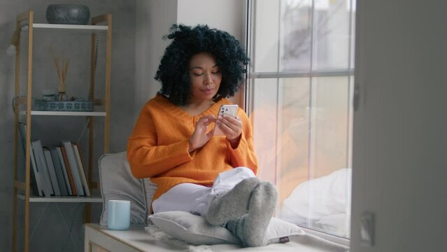 Beautiful curly hair woman using smartphone technology shopping online sale in slow motion. Portrait African American woman holding mobile phone watching social media, sits at window cold winter day. 