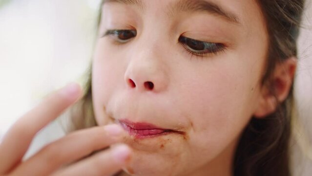 Closeup, girl child and licking fingers with chocolate, food or sweets in home for dessert, happiness and eating. Young female kid, lick hands and candy in family home in zoom of face in Los Angeles