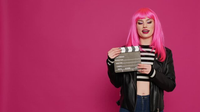 Beautiful cool girl showing filming slate on studio camera, working to record video footage or film montage on set. Glamour trendy adult saying action and recording, stylish punk filmmaker.