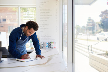 Male entrepreneur writing in book on checkout counter at coffee shop