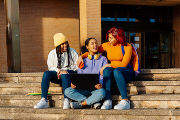group of three young latina girls sitting on the stairs outside the high school looking at a laptop...