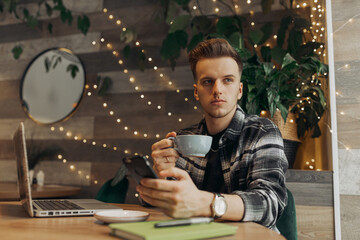 Serious young businessman having coffee break in cafe and browsing mobile phone while looking away with confidence 