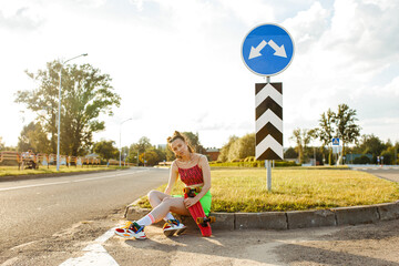 Cool young teenage female skater in vivid hipster outfit sitting with skateboard on road in summer...